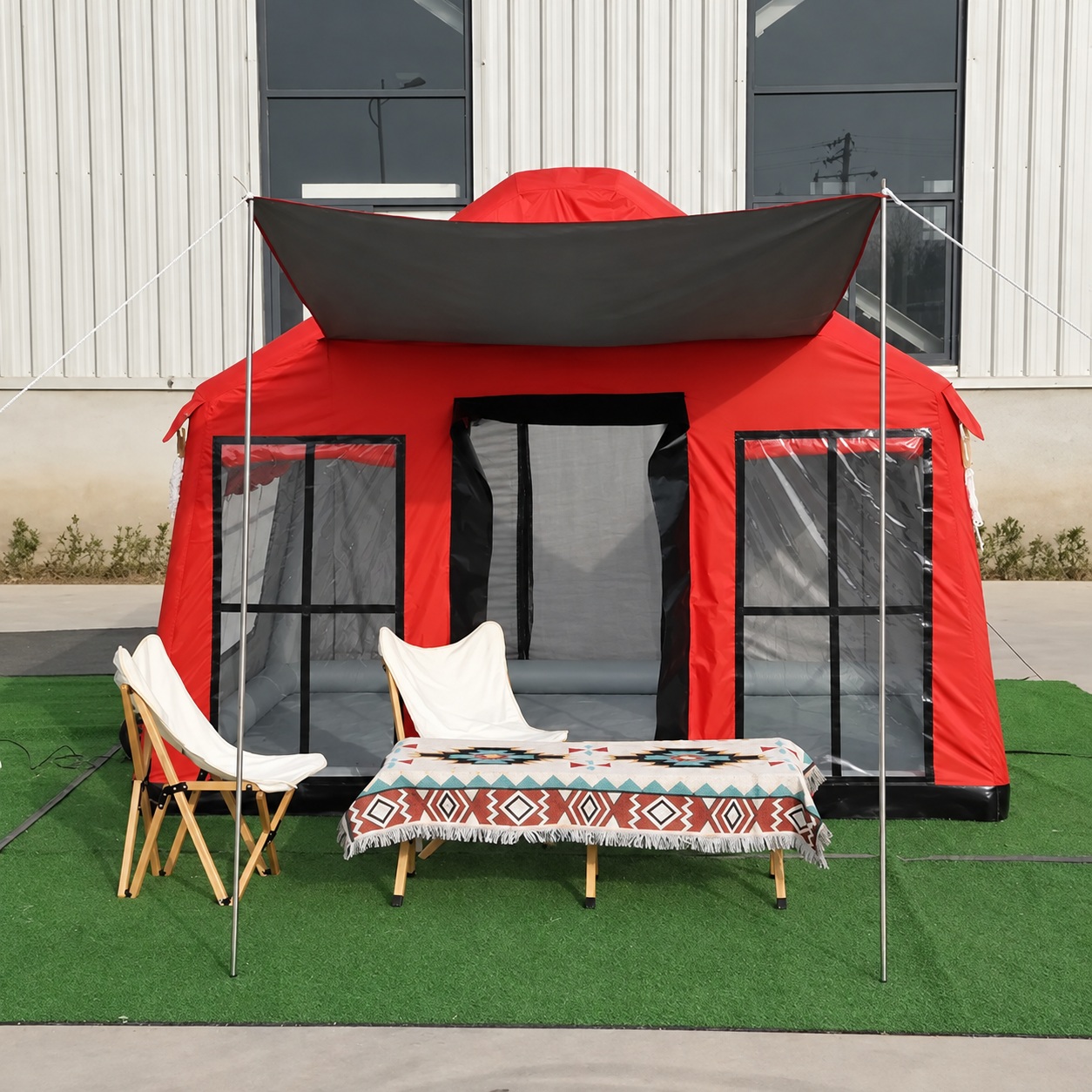 Red cabin tent with a raised front awning, dark-trimmed clear windows and an open doorway, shown with chairs and a table